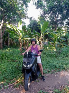 Tropikal ormanda, orman yolunda siyah scooterlı bir adam. Bali 'nin yeşil ormanında motosikletli bir adam. İnanılmaz bir doğa.