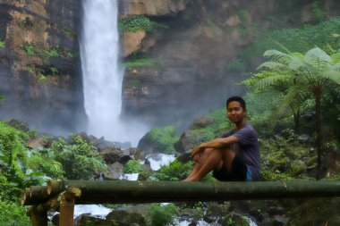 Genç bir Asyalı ormanda bir şelalenin yanında bambu bir bankta oturuyor.