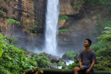 Yakışıklı Asyalı genç adam, güneşli bir günde bambu bankta oturmuş şelalede suya bakıyor. Metin için boşluk