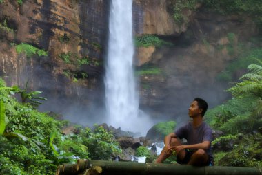 Yakışıklı Asyalı genç adam, güneşli bir günde bambu bankta oturmuş şelalede suya bakıyor. Metin için boşluk
