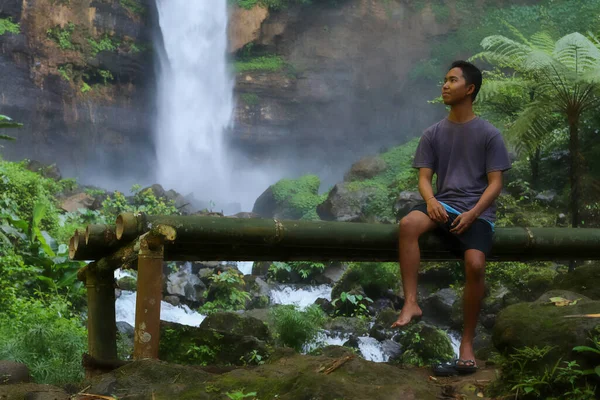 Молодий азіат сидить на бамбуковій лавці біля водоспаду в джунглях. — стокове фото