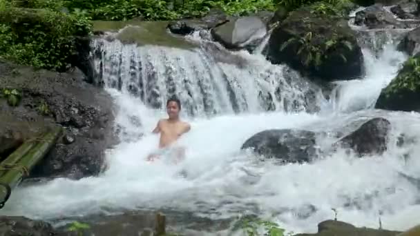 Man gets out of the cold water by a beautiful waterfall in tropical forest during sunny day - static shot motion — Stock Video