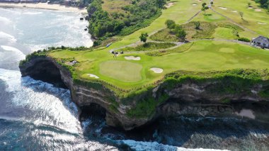Aerial shot of beautiful golf course on overgrown rocky cliff top in sunny luxury tourist resort on paradise island Bali, Indonesia. People swimming, surfers paddling in crystal clear blue ocean clipart