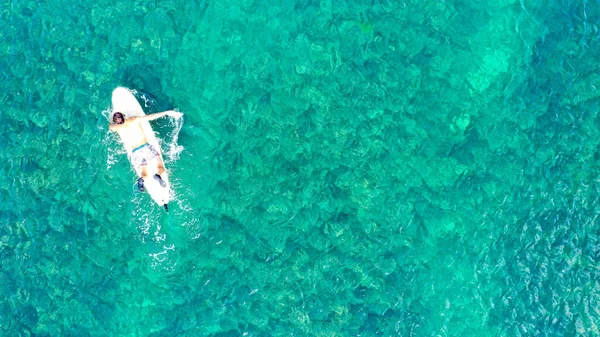 Vista aerea di giovani surfisti maschi galleggia su tranquille onde azzurre trasparenti sullo sfondo di una spiaggia accogliente con lettini e turisti in una giornata estiva soleggiata. Concetto di surf e vacanza Paradise — Foto Stock