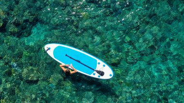 Zinde bir kadının havadan drone 'la çekilmiş fotoğrafı bir SUP tahtasının yanında denizde gevşiyor. Tropikal egzotik turkuaz açık denizde Stand Up Paddle tahtasıyla denizde bir kız.