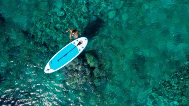 Zinde bir kadının havadan drone 'la çekilmiş fotoğrafı bir SUP tahtasının yanında denizde gevşiyor. Tropikal egzotik turkuaz açık denizde Stand Up Paddle tahtasıyla denizde bir kız.