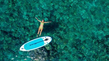 Zinde bir kadının havadan drone 'la çekilmiş fotoğrafı bir SUP tahtasının yanında denizde gevşiyor. Tropikal egzotik turkuaz açık denizde Stand Up Paddle tahtasıyla denizde bir kız.
