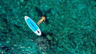 Zinde bir kadının havadan drone 'la çekilmiş fotoğrafı bir SUP tahtasının yanında denizde gevşiyor. Tropikal egzotik turkuaz açık denizde Stand Up Paddle tahtasıyla denizde bir kız.