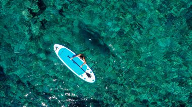 Zinde bir kadının havadan drone 'la çekilmiş fotoğrafı bir SUP tahtasının yanında denizde gevşiyor. Tropikal egzotik turkuaz açık denizde Stand Up Paddle tahtasıyla denizde bir kız.