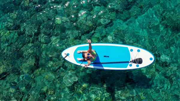 Mujer de arriba hacia abajo arrodillada en el tablero de SUP y mirando hacia arriba. Drone disparo de chica joven de pie remando en vacaciones — Foto de Stock