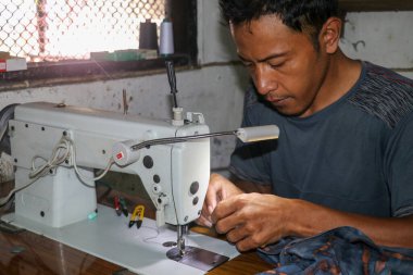 Asyalı adam dikiş makinesine elbise dikiyor. Orta yaşlı erkek terzi atölyede dikiş makinesi ve kumaşla çalışıyor.
