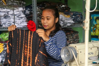 Genç bayan terzi ya da moda tasarımcısı elinde yeni Batik elbisesiyle dikiş makinesinin yanında oturuyor. Yerel giyim sektörünün odaklanmış kadın sahibi yeni dikilmiş elbiseyi kontrol ediyor