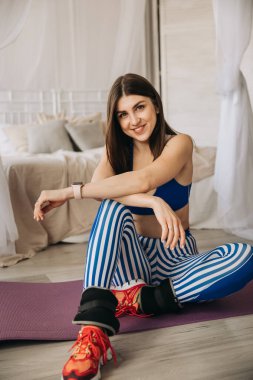 Mutlu sportif ve gülümseyen genç kız evde yoga minderinde oturuyor. Evde spor yapıyor, esneme hareketleri yapıyor..