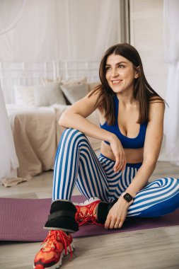 Mutlu sportif ve gülümseyen genç kız evde yoga minderinde oturuyor. Evde spor yapıyor, esneme hareketleri yapıyor..