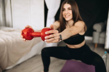 Siyah şık bir spor bluzu ve taytlı, ısrarcı ve kararlı bir esmer kadın, kollarını uzatmış halterlerle derin çömelmeler yapar..