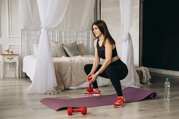 Israrcı ve kararlı siyah spor bluzu ve taytlı güzel bir esmer kadın, elinde dambıllarla derin çömelmeler yapar..