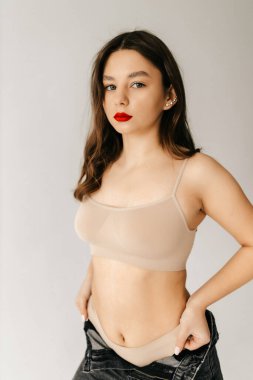 Stüdyoda model kız üstte ve pantolonla poz veriyor ve dudaklarında kırmızı ruj var.. 