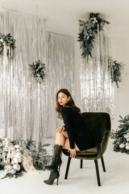 Siyah elbiseli manken beyaz stüdyoda sandalyede poz veriyor.