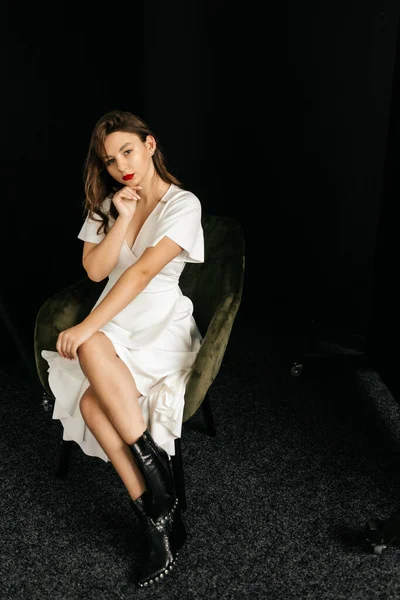 Beautiful young girl in a white dress sits on a chair on a black background — Stock Photo, Image