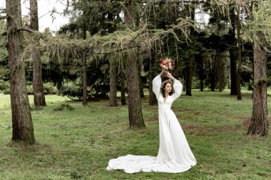 Beyaz gelinlik içinde güzel bir gelin ve yeşil bir ormanda elinde buketle.