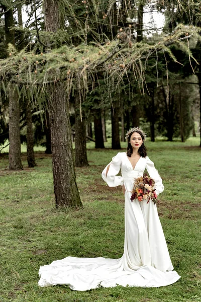 Beautiful bride in a white dress and with a bouquet in her hands on the background of the forest — Fotografia de Stock