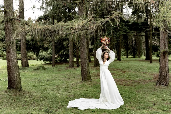 Beyaz gelinlik içinde güzel bir gelin ve yeşil bir ormanda elinde buketle.