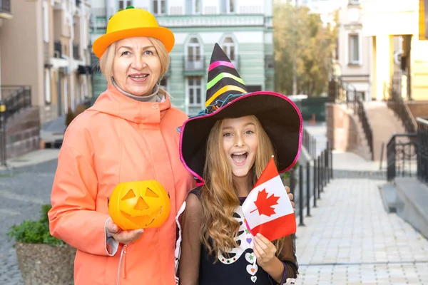 Grandmother and granddaughter with Canada flag and pumpkin, on the day of Halloween. Little girl in a witch costume.