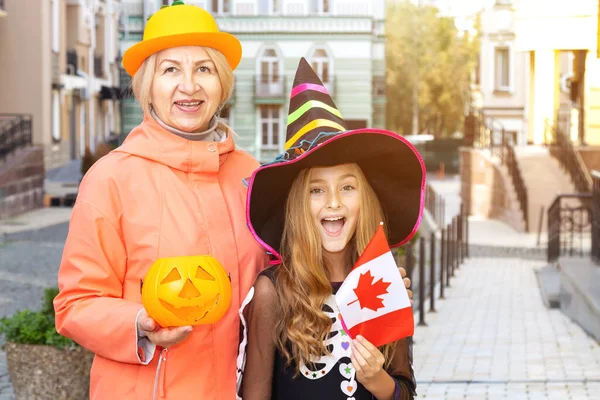 Grandmother and granddaughter with Canada flag and pumpkin, on the day of Halloween. Little girl in a witch costume.