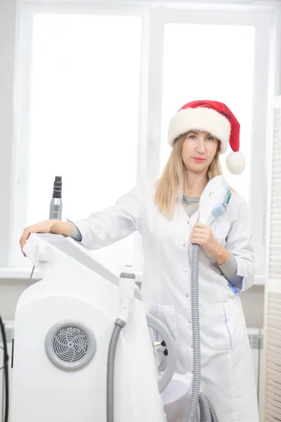 Retrato de una hermosa cosmetóloga de pie en una oficina de cosmetología con una herramienta de rejuvenecimiento de fotos en sus manos y un sombrero de Santa Claus listo para celebrar la Navidad y el Año Nuevo y recibir — Foto de Stock