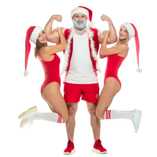 회색 턱수염을 기르고 산타클로스 복장을 한 건장 한 운동 선수가 붉은 수영복을 입고 하얀 배경에 고립된 채팔에 모자를 쓰고 섹시 한 두 소녀를 들고 크리스마스 사진을 찍기 위해 포즈를 취한다. — 스톡 사진
