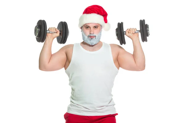 Un gran atleta atlético entrena bíceps levantando pesas de pie en un sombrero de Santa Claus listo para celebrar la Navidad aislado sobre un fondo blanco — Foto de Stock