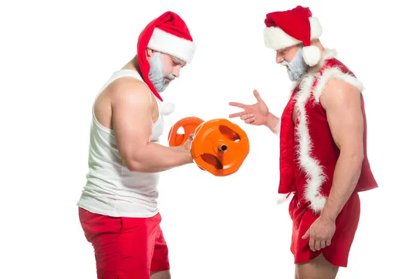 Vista lateral del musculoso Santa Claus en un sombrero y con una barba gris entrena a otro atleta en un disfraz de Navidad levantando una pesada barra aislada sobre fondo blanco — Foto de Stock