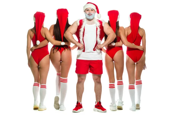 크리스마스. 티셔츠와 빨간 반바지를 입은 산발적 인 회색 수염 산타클로스의 전체 사진은 날씬 한 소녀들의 뒷모습 과 엉덩이에 손을 얹고 서 있는 강한 이두근을 보여준다. — 스톡 사진