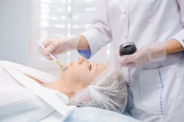 Побочный обзор рук косметолога, применяющего углеродную наногель на лицо молодой женщины с помощью кисти, готовящегося к процедурам углеродного пилинга — стоковое фото