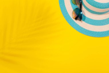 Sarı arkaplanda plaj aksesuarları, güneş gözlüğü ve palmiye yaprağı gölgesinde çizgili mavi şapka. Plaj tatili kavramı, deniz turu, sıcak güneşli yaz. Reklam alanı