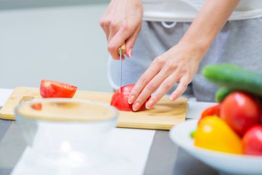 Vejetaryen vitaminli sebze salatası için kesme tahtasındaki bıçakla domatesleri doğrayan ve mutfakta kahvaltı hazırlarken cam bir kaseye koyan bir kızın ellerini kapat.