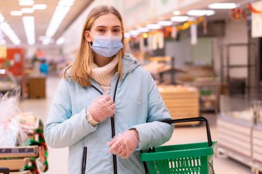 Bir süpermarkette elinde alışveriş sepetiyle tıbbi maske ve eldivenlerle duran genç ve güzel bir kızın ön görüntüsü. Karantina sırasında alışverişin yayılmasını önlemek için kurallara uygun olarak yapılması