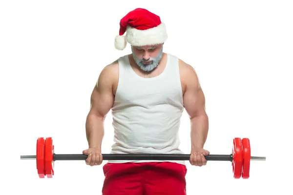 Święta. Silny umięśniony Święty Mikołaj z szarą brodą w świątecznym kapeluszu i czerwonych spodenkach podnosi ciężką sztangę na siłowni wykonując trening na białym tle — Zdjęcie stockowe