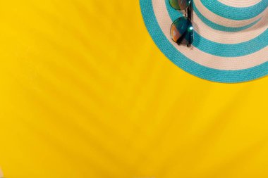 Sarı arkaplanda plaj aksesuarları, güneş gözlüğü ve palmiye yaprağı gölgesinde çizgili mavi şapka. Plaj tatili kavramı, deniz turu, sıcak güneşli yaz. Reklam alanı