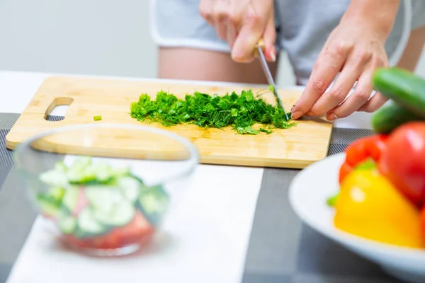 Primer plano de una chica manos de corte de vegetación con un cuchillo en una tabla de cortar para una ensalada de verduras de vitaminas veganas y ponerlos en un recipiente de vidrio mientras se cocina el desayuno en la cocina — Foto de Stock