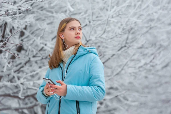 Elöl egy gyönyörű tinédzser lány zenét hallgat fejhallgatóval egy okostelefonon, miközben sétál egy havas téli parkban, félrenéz. — Stock Fotó