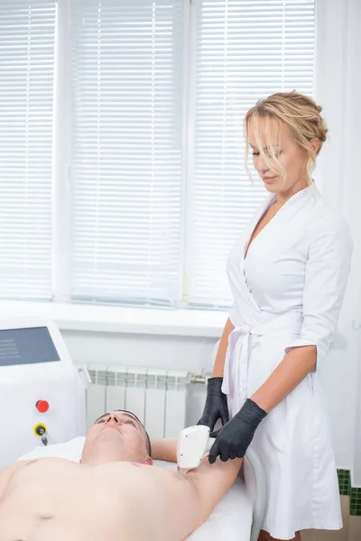 Um belo cosmetologista no trabalho em seu escritório de cosmetologia moderna durante o procedimento de depilação a laser da axila de um jovem — Fotografia de Stock
