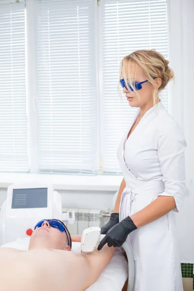 Eine hübsche Kosmetikerin und ihre Kundin in blauer Schutzbrille bei der Laser-Haarentfernung aus der Achselhöhle eines jungen Mannes — Stockfoto