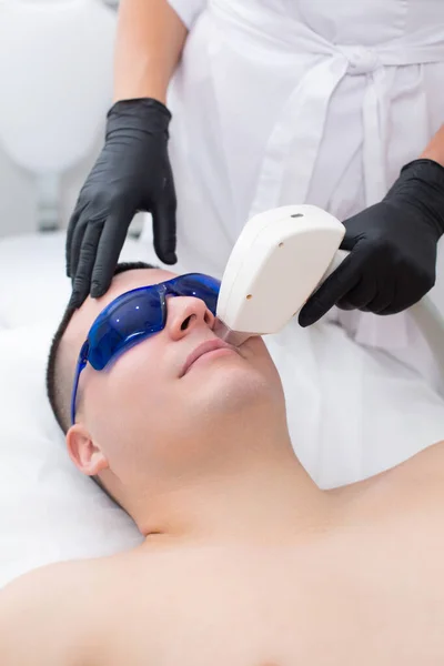 Close-up do rosto de um jovem bonito em óculos de segurança azuis durante um procedimento de depilação a laser em seu rosto — Fotografia de Stock