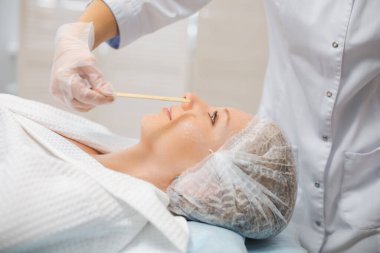 Kozmetik uzmanının elleri yüz maskesi takıyor, çubuk kullanıyor, deriyi sulandırıyor ve yüzü parlıyor. Çekici bir kadın gülümseyerek rahatlıyor ve spa prosedürlerinden zevk alıyor.