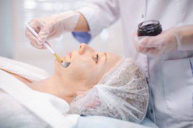 Genç hasta yüzüne karbon nanojel uygulayan kozmetoloji uzmanının elleri, fırça kullanarak karbon soyma prosedürlerine hazırlanıyor.