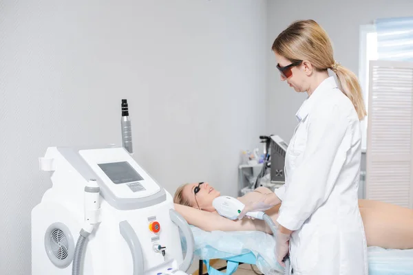 Foto av en modern kosmetologi anordning och en kvinnlig hudläkare i skyddsglasögon utför ett förfarande för laser epilering av armhålor av en ung flicka — Stockfoto