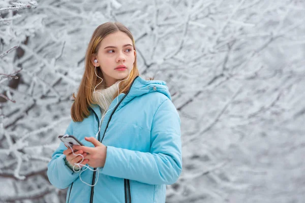 Вид спереду красива дівчина-підліток слухає музику з навушниками в смартфоні під час прогулянки в засніженому зимовому парку — стокове фото
