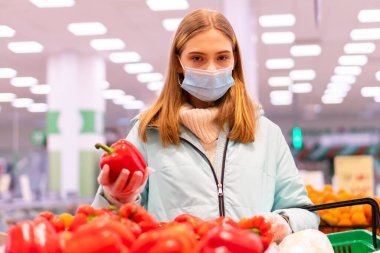 Tıbbi maskeli ve eldivenli genç bir kızın süpermarkette dolmalık biber seçerken çekilmiş fotoğrafı. Koronavirüs salgını sırasındaki anti-salgın önlemlerine uygun olarak alışveriş.