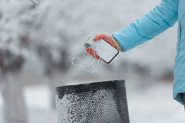 Zbliżenie młodej kobiety zamarzniętej dłoni wyrzucającej zepsuty smartfon i słuchawki do kosza podczas spaceru po śnieżnym parku zimowym — Zdjęcie stockowe